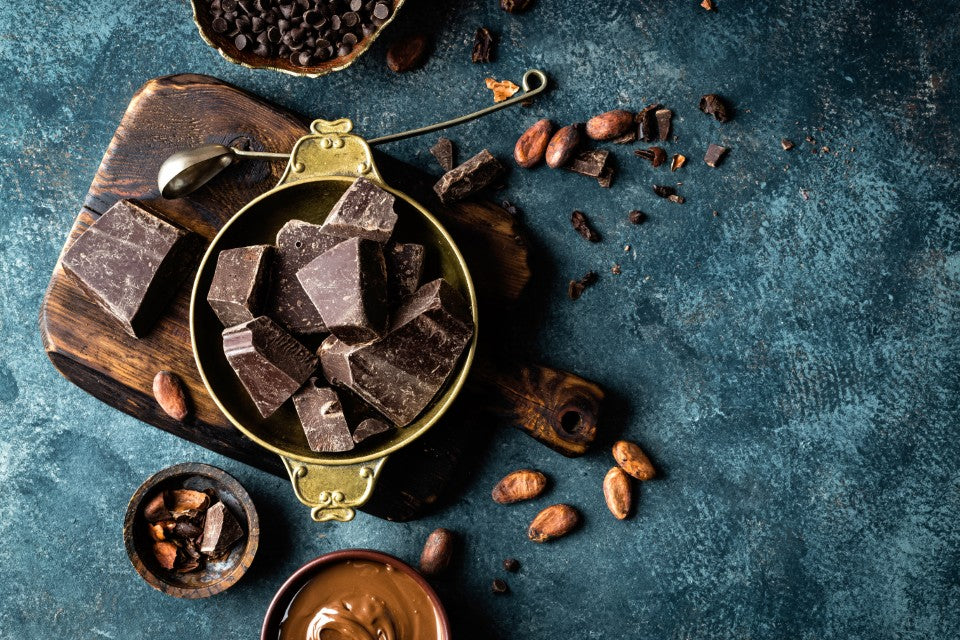 Dark chocolate chunks in a brass bowl with cocoa beans, cocoa nibs and cocoa powder on a blue-grey surface