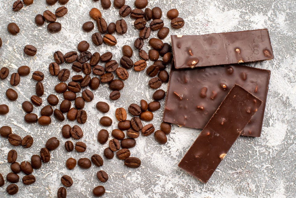 Dark chocolate bars with nuts next to scattered coffee beans on a light grey surface